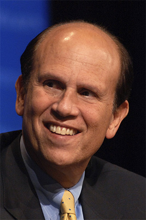 A headshot photo of Michael Milken smiling, wearing a grey suit, blue shirt, and yellow tie.