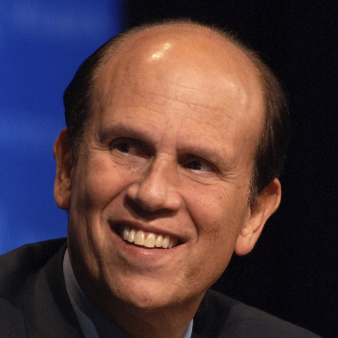 A headshot photo of Michael Milken smiling, wearing a grey suit, blue shirt, and yellow tie.